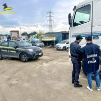 Autorità e mezzi della Guardia di Finanza in un'area di deposito veicoli: agenti parlano con due persone accanto a camion e auto ufficiali.
