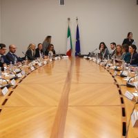Cerimonia di vertice in una sala riunioni, uomini e donne seduti attorno a un grande tavolo, bandiere allashire sullo sfondo.