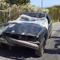 Automobile d'epoca rovinata su strada, telo parzialmente copre il parabrezza, contorni danneggiati, sfondo giardino e recinzione.