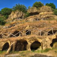 Scogliera rocciosa con fessure, grotte arancioni e passerelle in legno, circondate da vegetazione verde e cielo azzurro.