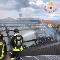 Parcheggio di un edificio in fiamme con vigili del fuoco che spruzzano acqua, fumo e strutture carbonizzate sul tetto.
