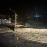 SCENA NOTTURNA: strada fatiscente con cordone di luci stradali, marciapiede sbrecciato e pozze d’acqua riflettenti.