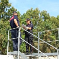 Due ufficiali di polizia in uniforme discutono su una passerella metallica, contornati da alberi e cielo azzurro.