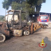 Automezzo da cantiere danneggiato con attrezzature metalliche arrugginite, veicoli fermi su terreno terroso, presenza di camion rosso sullo sfondo.
