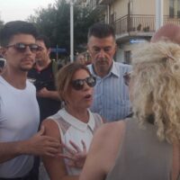 Gruppo di persone in strada, primo piano di una donna con occhiali e mani alzate, circondati da altri individui, atmosfera di conversazione.