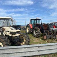 Trattori agricoli fermi all’aperto su terreno erboso, con cielo azzurro e nuvole, attrezzature e trattore rosso in primo piano.