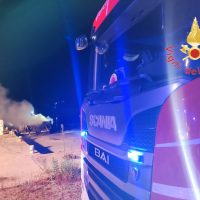 Auto dei pompieri al lavoro, autobotte blu con sirene accese, fumo al percorso e fuoco all’orizzonte su strada desertica.