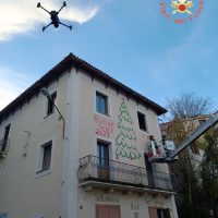 Kontrastata casa gialla con decorazioni natalizie, persona in salto dal tetto, cielo azzurro e albero sullo sfondo, atmosfera felice.