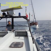 Barca della Guardia Costiera in mare aperto, rotta verso sinistra, equipaggio in pronto intervento, testo giallo in alto a sinistra.