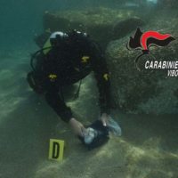 Immersione subacquea: subacqueo con attrezzatura estrae un oggetto dal fondale, segnalini numerici sul selciato e logo Carabinieri IBO.