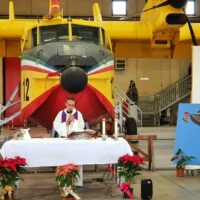 Prete che celebra una messa all'aperto davanti a un aereo giallo, con altarare bianco e decorazioni floreali rosse e verdi.