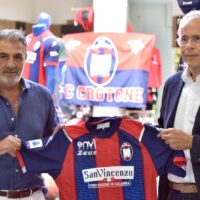 Due uomini posano con una maglia da calcio rossoblù, decorata con sponsor e una bandiera del club sullo sfondo.