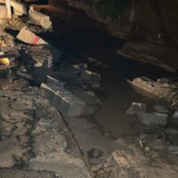 Ruderi e detriti lungo una strada sporca: copertoni e calcestruzzo rotti, pozzanghera d’acqua scura, atmosfera urbana degradata.
