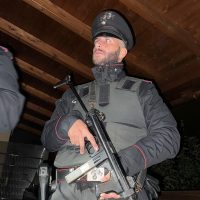 Uomo in divisa militare nera con berretto, equipaggiamento tattico e fucile, in interno legno.