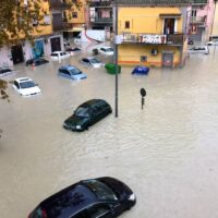 Risaia città allagata con auto e persone sulla strada sommersa, edifici colorati sullo sfondo e luci stradali visibili.