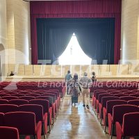 Teatro interno con sipario rosso e platea vuota; due persone camminano lungo il corridoio verso la scena illuminata.