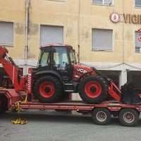 Trattore fuoristrada rosso su carrello, caricatore anteriore in uso, sullo sfondo edificio giallo con finestre e vetrine.