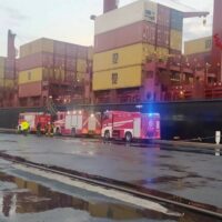 Immagine di un incendio in un deposito: autoscala, veicoli dei vigili del fuoco, fiamme visibili tra container impilati, strada allagata.