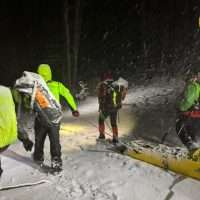 Escursionisti in alta quota durante una tempesta di neve, indossano giacche fluorescenti e zaini, camminando tra alberi coperti di neve.