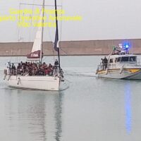 Due catamarani da regata in un porto: equipaggi sulla passerella, acqua calma e murata sullo sfondo, atmosfera di vento e navigazione.