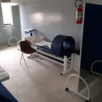 Sala medica con policy: attrezzature su banco, sedie, scaffale e dispositivi di analisi, luci bianche, ambiente clinico ordinato.