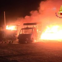 Veicolo in fiamme all’aperto durante un incendio notturno, fuoco intenso e fumo rosso, contesto urbano rurale.