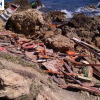Scoglio roccioso lungo la costa con tavole di legno rotte e detriti su spiaggia rocciosa, mare azzurro sullo sfondo.