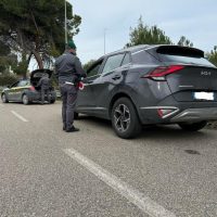Ragazzo fermo vicino a una moto e un’auto scura su una strada cittadina, controlli di sicurezza o verifica documenti.
