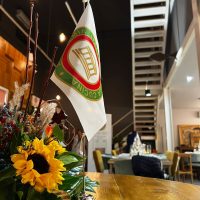Interno accogliente di ristorante, tavolo con bouquet di girasole e bandiera verde-rossa sullo sfondo, atmosfera luminosa e confortevole.