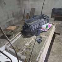 Lavandino esterno sporco e attrezzi arrugginiti in un cortile grigio, con tappeto e oggetti vari sparsi su una superficie allungata.