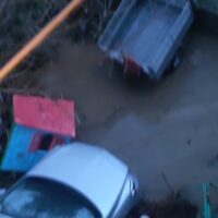 Vista aerea di auto parcheggiate e veicoli immersi in una strada allagata, con riflessi d’acqua e macchie scure sul manto stradale.