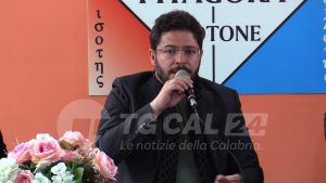 Uomo con barba al microfono durante conferenza, sfondo colorato con loghi e fiori sul tavolo