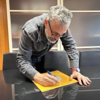 Uomo maturo con occhiali piegato sul tavolo scrive su foglio giallo, in un ambiente d'ufficio con mobili moderni e pareti chiare.