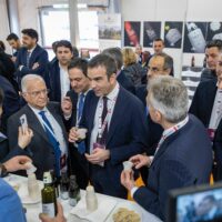 Gruppo di uomini in abiti formali brindano e conversano durante un evento di networking, con tavolo di degustazione di vini e cibi sullo sfondo.