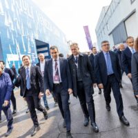 Uomini d'affari vestiti elegantemente camminano all'aperto in un cortile commerciale con edifici moderni e cartelloni sullo sfondo