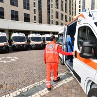 Operatore sanitario in divisa arancione che entra in un'ambulanza, sfondo di altre ambulanze e edifici, contesto urbano.