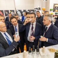 Gruppo di uomini in un evento aziendale brindano con bicchieri, in abiti formali, davanti a una tavola con bevande e snack.