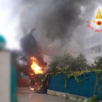 Immagine di un incendio all'aperto tra edifici: fiamme ardenti, fumo denso e persone in pericolo vicino a una recinzione blu.