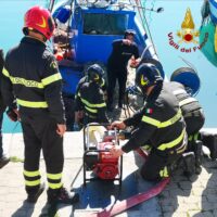 Soccorritori in attrezzatura antincendio assistono durante un intervento su una barca, usando una pompa e una sacca di emergenza su banchina soleggiata.