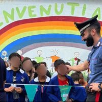 Gruppo di bambini in uniforme davanti a un murale arcobaleno con la parola Benvenuti; presenza di un conducente in divisa che li guida.