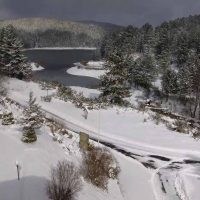 Paesaggio invernale: laguna ghiacciata tra alberi innevati e colline, cielo grigio, neve fresca e con piccoli sentieri innevati.