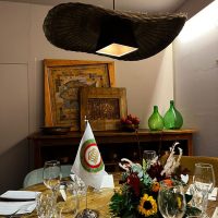 Tavolo apparecchiato per una cena elegante con fiori colorati, bicchieri e piatti, vicino a un quadro e lampada pendente in ambiente urbano.