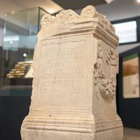 Slab tombstone in pietra chiara con rilievi scolpiti, esposta in una mostra museale moderna.
