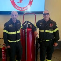 Due vigili del fuoco in uniforme accanto a estintori rossi, in laboratorio o centrale, sorridenti e pronti all’intervento.