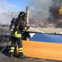 Pompiere in azione con autorespiratore, fronteggia un incendio al di là di una barriera arancione, fiamme e fumo sullo sfondo.