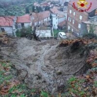Veduta di una frana su un terreno roccioso, con abitazioni sullo sfondo e strati di terra e detriti in primo piano.