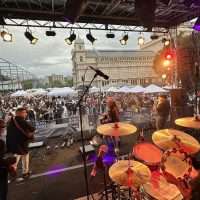 Concerto dal vivo su palco all'aperto con batteria e microfoni, pubblico in piazza e palco illuminato al tramonto.