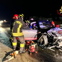 Incidente stradale notturno: vigili del fuoco e soccorritori attorno a un'auto frontalmente incidentata sospesa su una strada, attrezzi sul terreno.