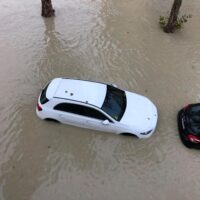 Auto bianca incastrata in una strada allagata tra alberi, riflessi sull’acqua marrone e veicolo nero laterale.