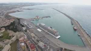 Veduta aerea di porto turistico con nave ormeggiata, edifici lungo la banchina e acque turchesi; paesaggio marino dominante.